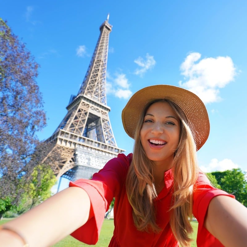 Young Blonde Woman Wearing A Red Summery Blouse And A Straw Hat As She Smiles Taking A Selfie With The Eiffel Tower In Paris, France, Europe
