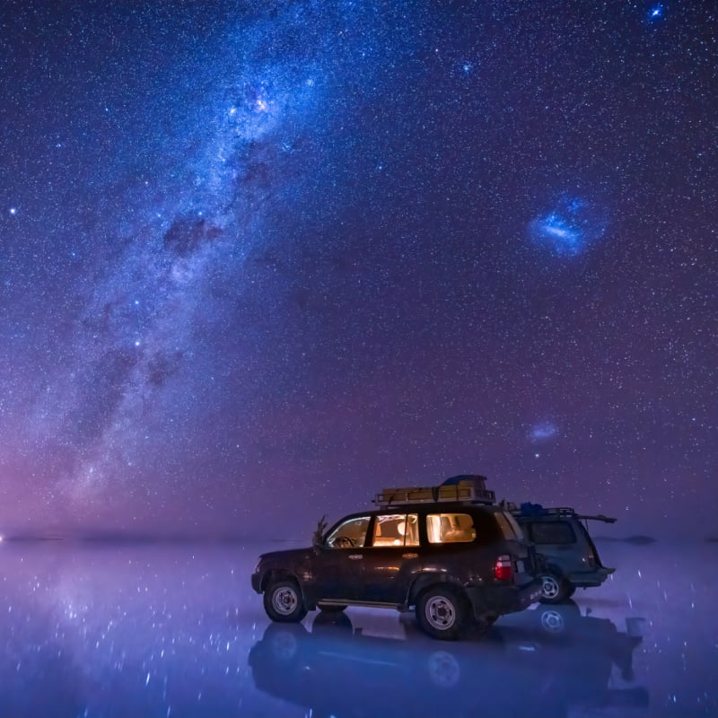 milky way and starry sky over the salt flat of Uyuni, Bolivia, with 2 SUVs parked on the reflective lake surface.
